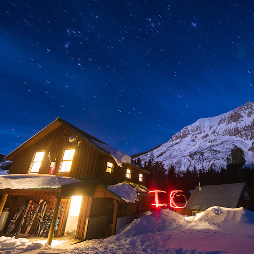 House with lights on in night with stars with mountain in background.